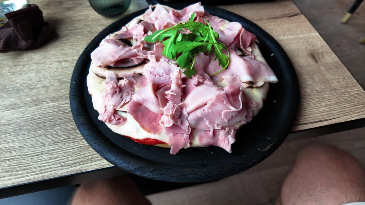 Freshly prepared pizza topped with slices of ham and arugula, served on a black plate over a wooden table in a cozy dining setting. Ideal for food and culinary themes