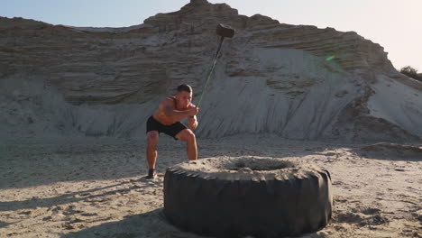 muscle athlete strongman man hits a hammer on a huge wheel in the sandy mountains in slow motion at sunset. the dust from the wheels rises.