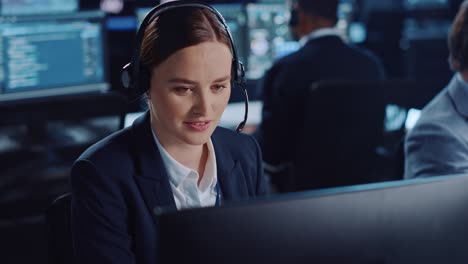 una bella specialista del supporto tecnico al cliente sta parlando con un auricolare mentre lavora su un computer in una stanza di monitoraggio e controllo buia piena di colleghi e schermi di visualizzazione.