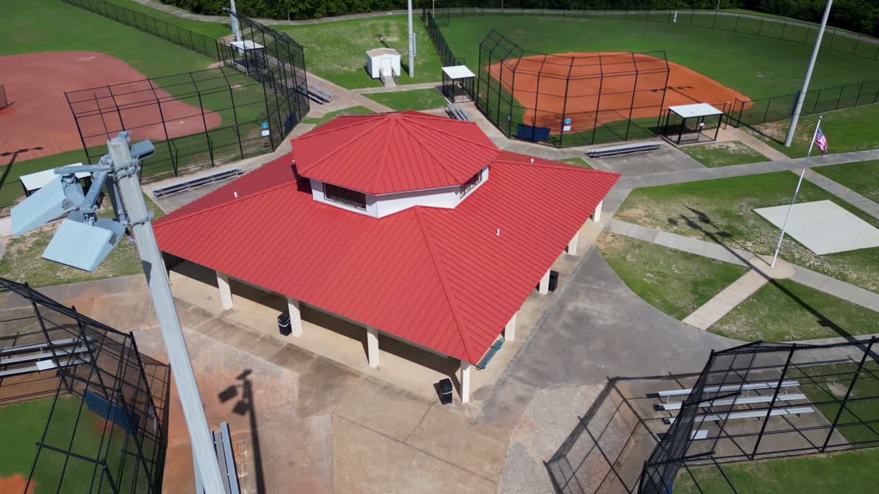 slowly pushing and tilting drone shot towards concession building between light poles and baseball fields at a small park.