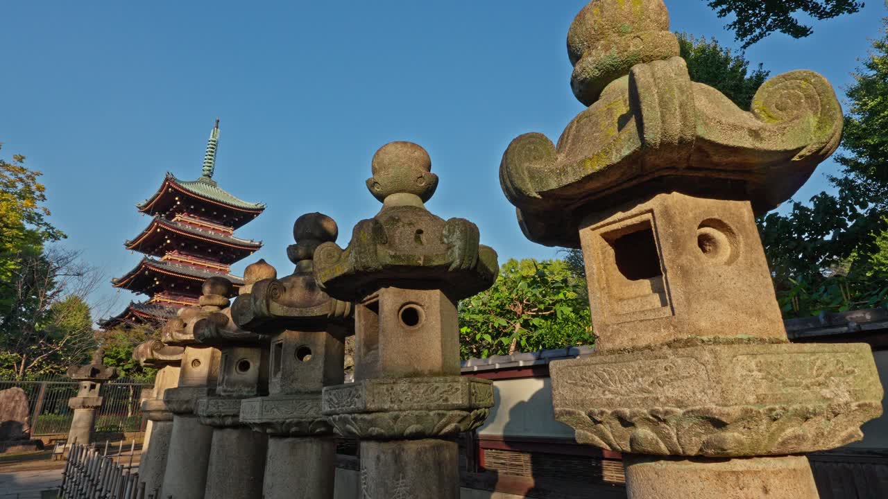 A traditional Japanese pagoda stands prominently behind a row of ancient stone lanterns in Ueno Park, Tokyo. The clear sky and serene setting highlight the timeless beauty of this historical landmark.