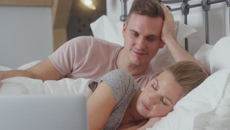 couple in bed wearing pyjamas streaming movie on laptop as woman sleeps