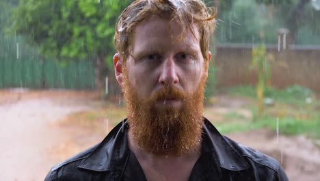 retrato en cámara lenta de un pelirrojo barbudo parado bajo la lluvia torrencial y empapándose mientras mira a la cámara