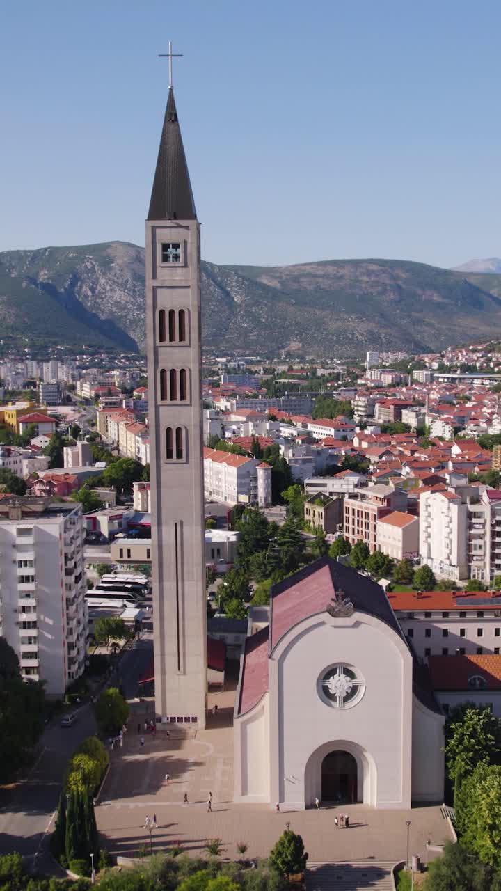 Aerial view of the Church of St. Peter and Paul with its prominent bell tower, surrounded by the urban landscape of Mostar, Bosnia and Herzegovina. Vertical Video, Orbit Motion