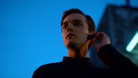 Young man adjusting an earbud against a blue sky