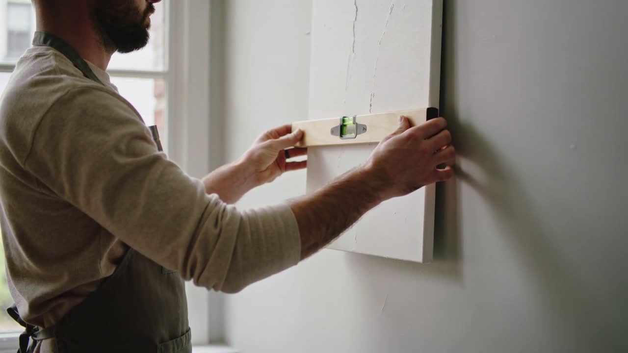 Placing wooden spirit level, bearded maker in dark apron aligning white panel on wall for marking