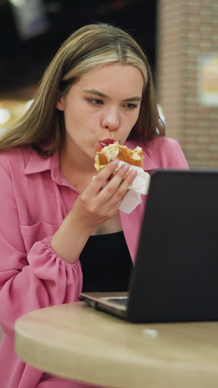 mujer en vestido rosa multitarea en un centro comercial ocupado, trabajando en su computadora portátil mientras come una hamburguesa, parece centrada en su trabajo, equilibrando la productividad y su comida en una mesa de madera