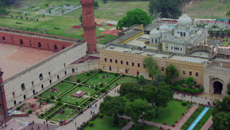 drone flight to the sikhism temple passing over a mosque, children and ladies waving at the drone, white temple and a red mosque, a beautiful park at the front of the mosque and temple