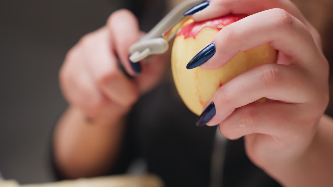 Close view of manicured hands peeling red apple with peeler in bright kitchen, blue nails visible, careful fruit prep for healthy snack, smooth controlled strokes, fresh produce focus