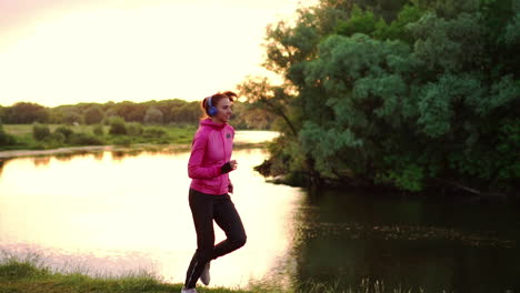 Brunette-with-long-hair-in-headphones-runs-along-the-river-in-the-Park-in-the-morning-at-sunrise-in-the-summer-in-a-pink-jacket-and-black-pants