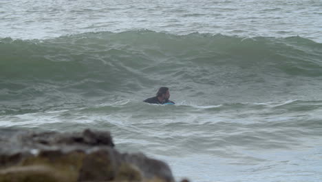 hombre deportivo en traje de neopreno con pierna artificial acostado en la tabla de surf y nadando en el océano cuando una ola lo cubre