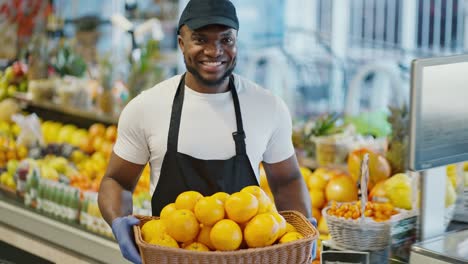 Portrait-of-a-happy-man-with-Black-skin-color-a-supermarket-worker-in-a-white-T-shirt,-black-cap-and-black-apron-holds-in-his-hands-a-large-basket-of-oranges-near-a-counter-with-citrus-fruits-and-berries