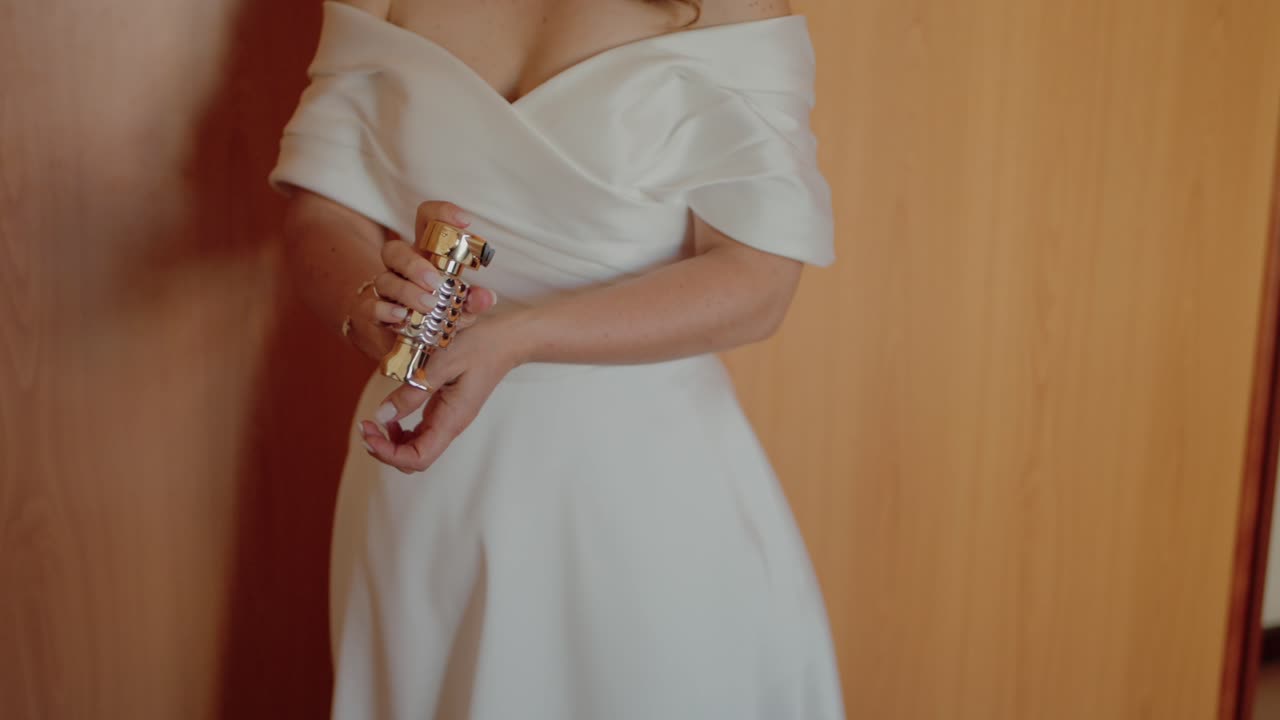 Bride in elegant white dress applying perfume on wrist against wooden wardrobe