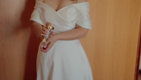 bride in elegant white dress applying perfume on wrist against wooden wardrobe