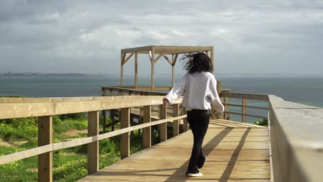 Young-woman-walks-briskly-and-happily-skipping-along-a-wooden-lookout-point-to-view-the-seascape-on-the-coast-of-Algarve,-Portugal