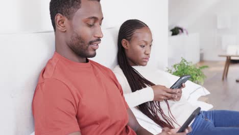 African-american-couple-sitting-up-in-bed,-both-using-smartphones,-in-slow-motion