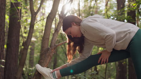 woman stretches leg on branch after jogging in forest. healthy lady in sportswear enjoys active lifestyle training on summer day surrounded by dense forest