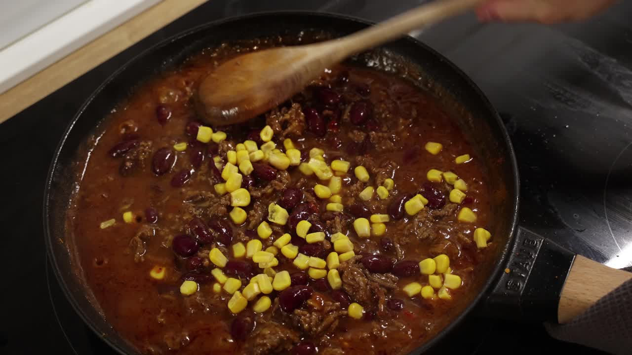 Molho de carne estilo chili sendo adicionado ao milho na panela, criando uma refeição caseira reconfortante.