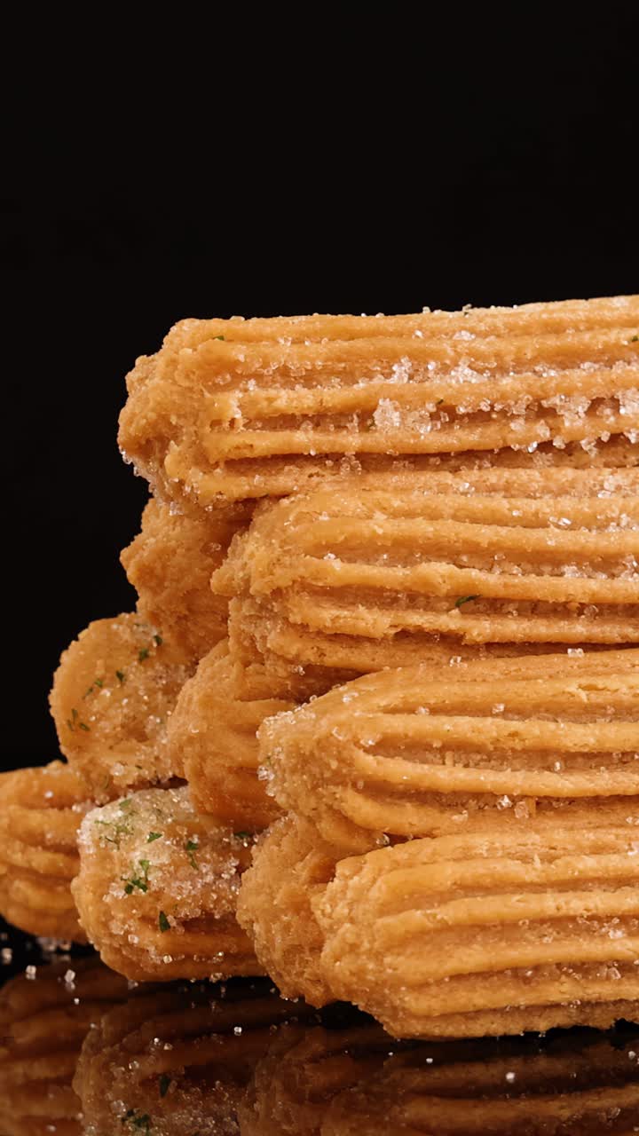 Golden churros coated in sugar rotate smoothly on a reflective black surface, illuminated by soft studio lighting, highlighting texture and detail throughout the sequence