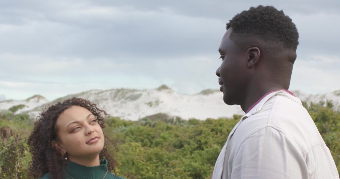 Couple se touchant les mains, la femme en pull vert tendant la main, l'homme l'étreignant sur des dunes pour la réconforter.