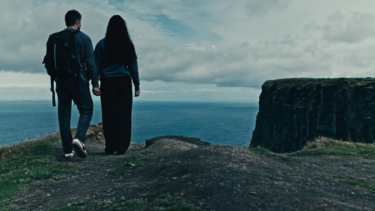 A couple stands hand in hand facing the cliffs and sea under a cloudy sky, wind blowing gently at the Moher coast - Ireland