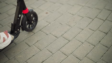 close-up of a woman's foot with painted nails, wearing white sandals, riding a black scooter on an interlocked pathway