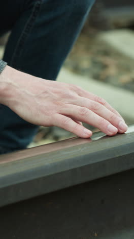 una vista de cerca de un hombre con un blazer gris y pantalones vaqueros azules arrodillado junto a una vía ferroviaria, tocando cuidadosamente la vía con la mano, luego retiró la mano de la vía del sendero, mirando su mano
