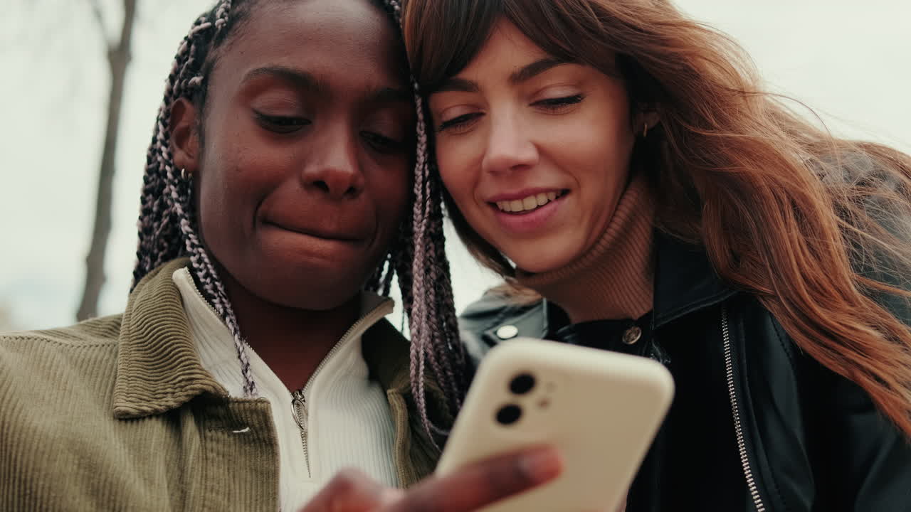 Due giovani donne di diverse etnie sorridono e condividono uno smartphone all'aperto