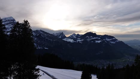 view of the swiss alps at sunset, featuring snow-covered slopes, silhouetted pine trees, and majestic mountain peaks under a dramatic, cloudy sky