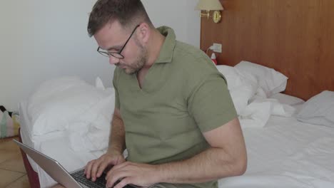 a man sits in a hotel room using his laptop. his frustrated expression suggests stress, annoyance, or working while on holiday instead of relaxing