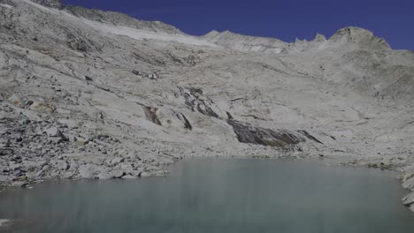 el dron sube sobre un lago glacial único con un río que fluye sobre piedras de granito, moldeado por el glaciar neves visible en el fondo