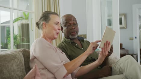 Glückliches-älteres,-Vielfältiges-Paar,-Das-Hemden-Trägt-Und-Im-Wohnzimmer-Ein-Tablet-Benutzt