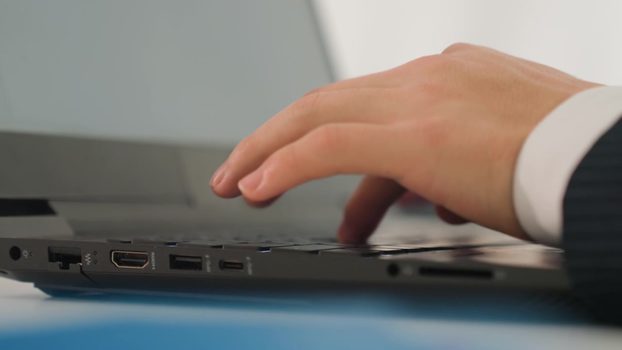 Mani caucasiche inquadrate dal basso che gestiscono un laptop su una scrivania blu, polsino della giacca visibile, digitazione rapida e reattiva durante una riunione, project manager che analizza i dati, responsabile delle vendite che prepara una presentazione, urgente.