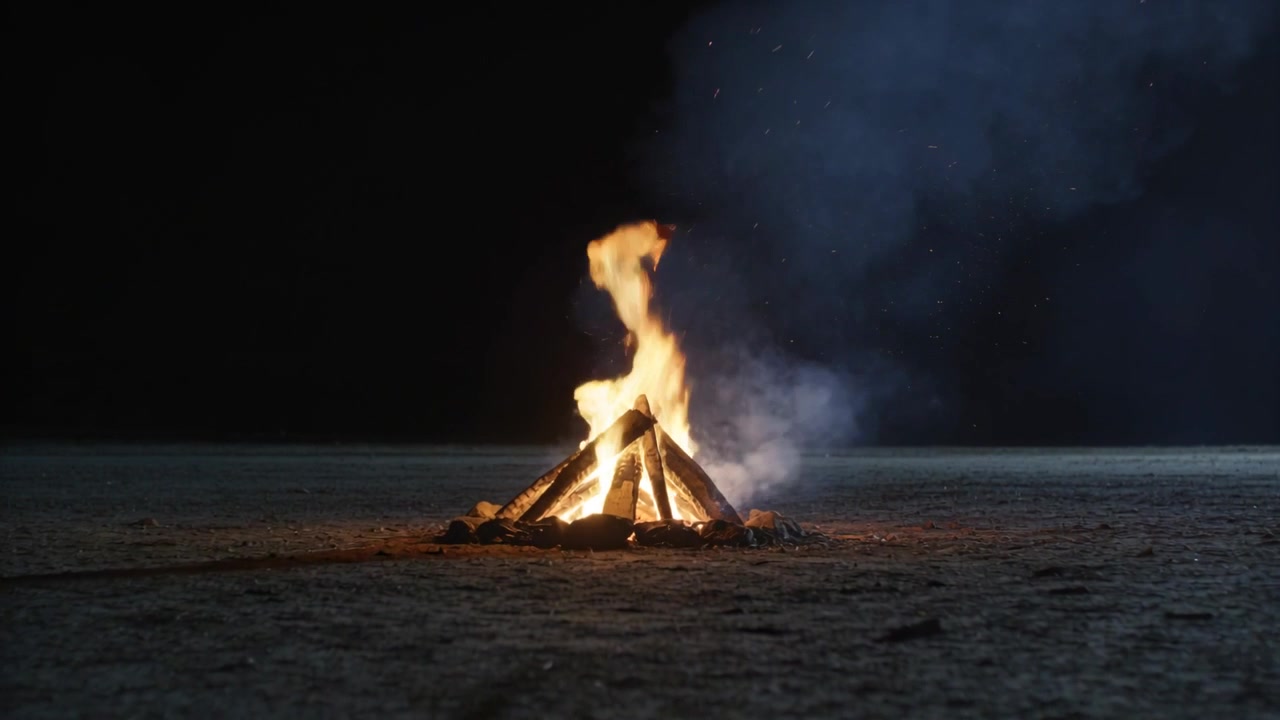 Braci che accendono la legna disposta a teepee, il falò che cresce, mandando fumo verso destra sulla sabbia piatta, illuminando le rocce.