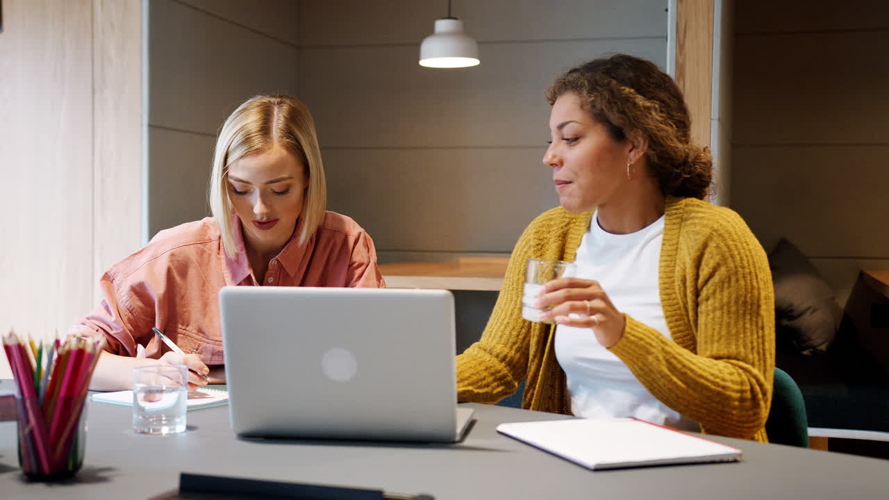 Front view of two young adult women working together sitting at a laptop computer in an office, selective focus