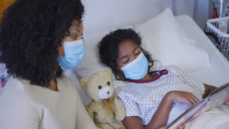 mixed race mother and daughter lying on hospital bed reading book, both wearing face masks