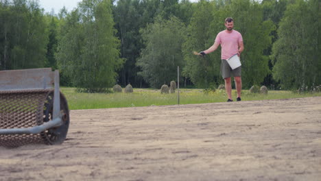 a man sowing grass on a field near a forest in the summer with a tool in the foreground