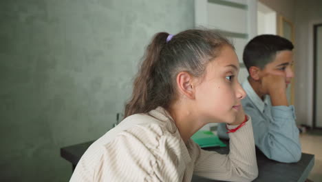 Thoughtful girl and bored boy sitting silently at table with green folders in modern room, both looking away, conveying lack of interest, school stress, emotional distance, and disconnection