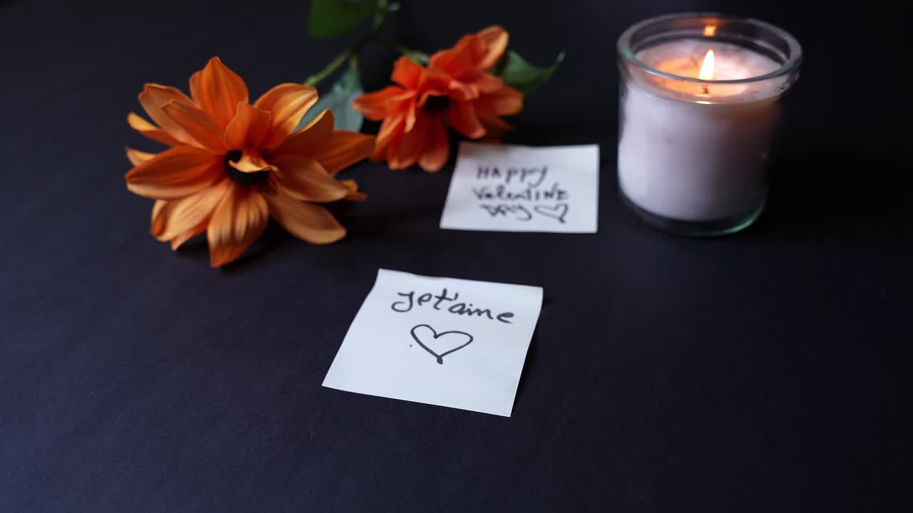 Close-up of a hand holding “Je t’aime” love note with heart beside a lit glass candle and orange flowers on a dark tabletop, warm candlelight creating an intimate Valentine’s mood