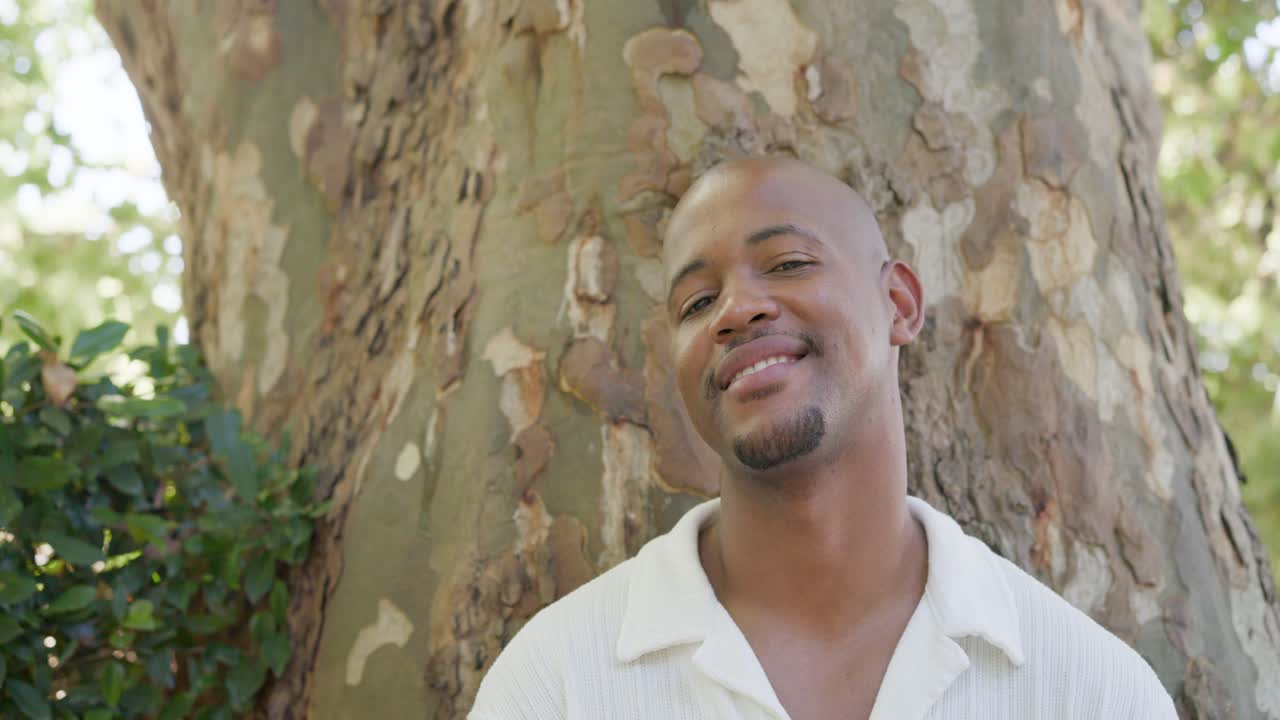 African American man leaning on right of tree wearing white shirt, reacting to sunlight and smiling