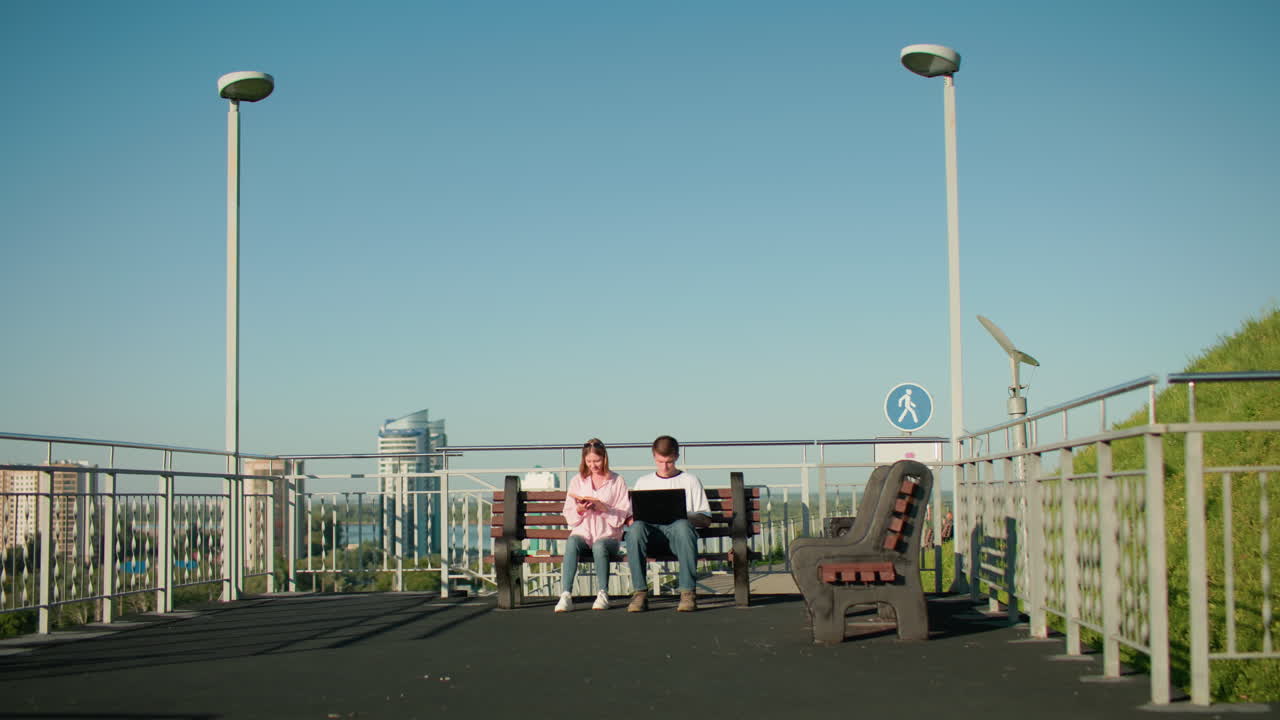 Two students seated outdoors on wooden benches, boy typing on laptop while girl points at her book, surrounded by urban cityscape, railings, greenery, and clear blue sky on a bright sunny day