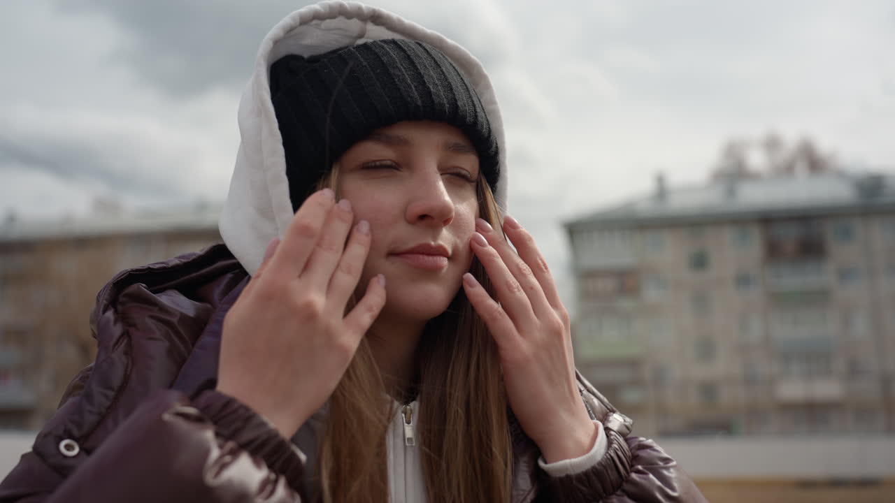 흰색 비니를 똑바로 쓰고 후드를 올리는 백인 여성, 갈색 코트와 레이어드된 후드가 보임, 쌀쌀하고 흐린 날씨에 산책을 가기 위한 은근한 준비, 솔직한 교외 거리 배경, 집중된 표정