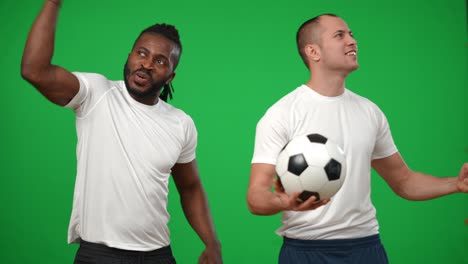 inspired satisfied african american and caucasian men cheering at green screen looking around. successful confident footballers rejoicing goal at chromakey background. sport and success concept.
