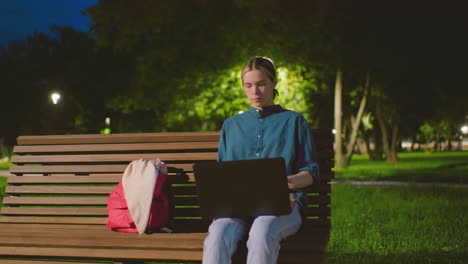 young lady seated on outdoor bench at night, with red bag nearby, opening laptop under soft light, background shows serene park with trees, light pole, lush greenery and deep blue sky