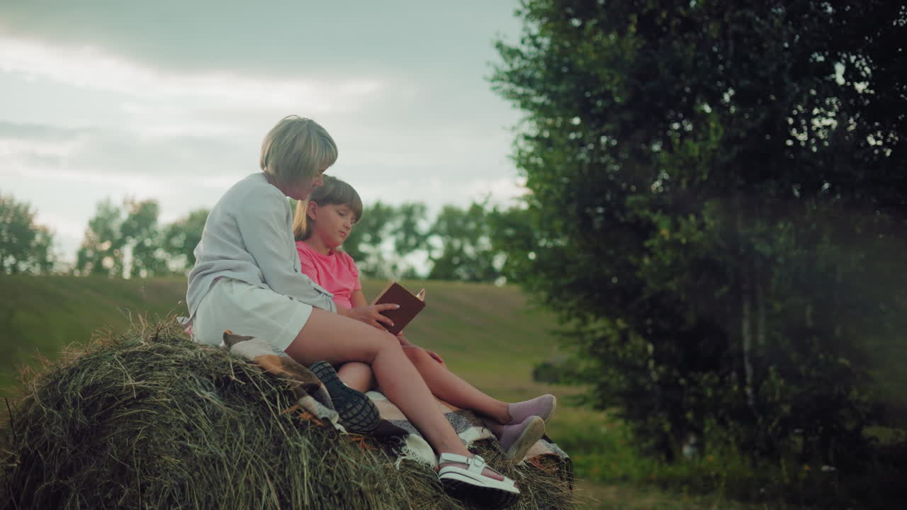 어머니가 딸을 안고 마른 잎자루 한 어리에 앉아 모자를 고 저녁에 열린 들판에서 책에 깊이 몰두하는 사랑스러운 순간.