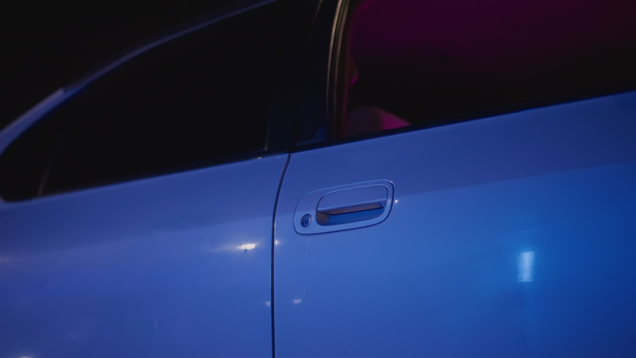 Puerta azul noche de un coche abierta por una mano furtiva, brillo rosa del asiento interior y luces del puente visibles, primer plano del asa y sutil destello de los faros, silueta de figura en vaqueros, lleno de suspense