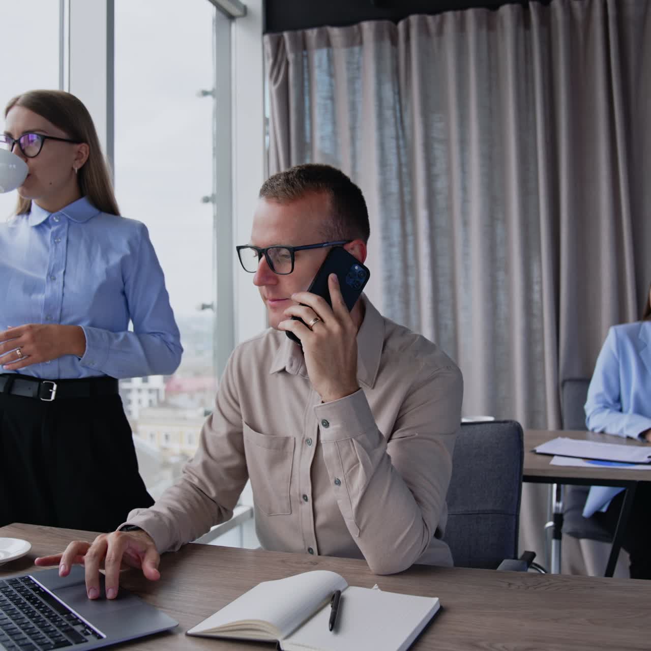 People working in office. Employees using phones and laptops at their work. Female colleague drinks coffee standing at window