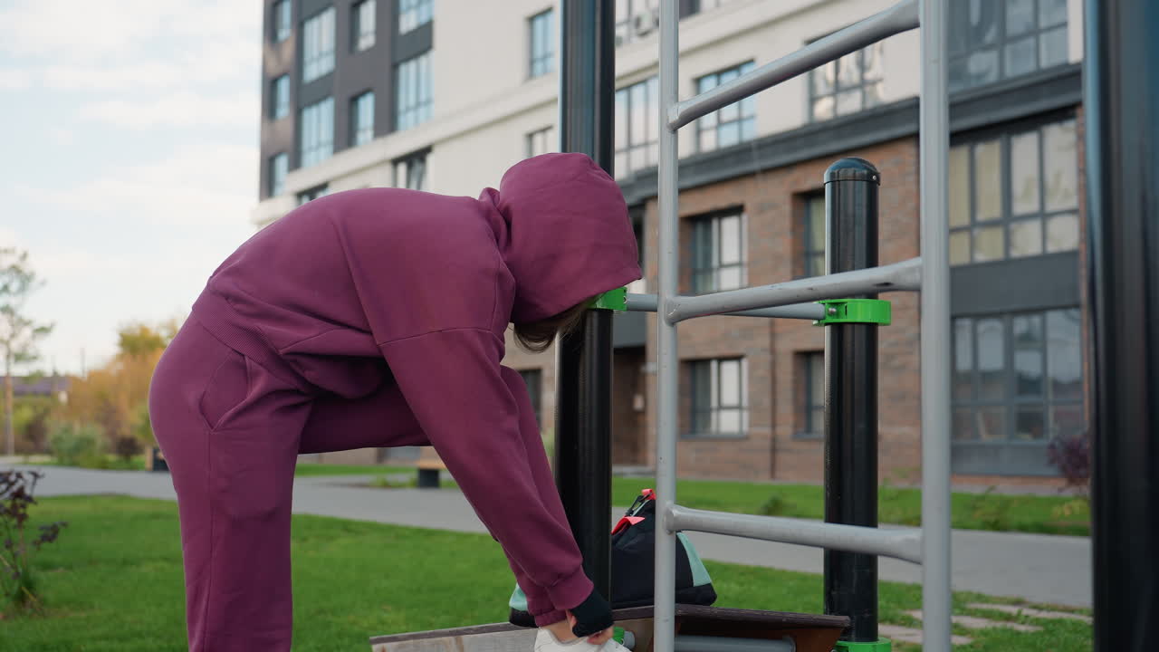 Determined woman walks toward iron bars places gym bag on corner then props foot to tie shoelace before workout in urban outdoor fitness park under clear sky near modern residential complex