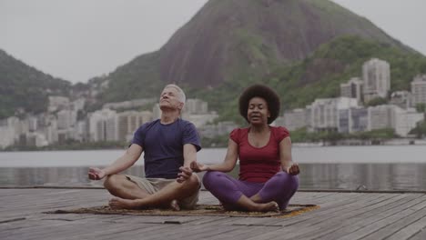 two people meditating outdoors by a lake with a city and mountains in the background