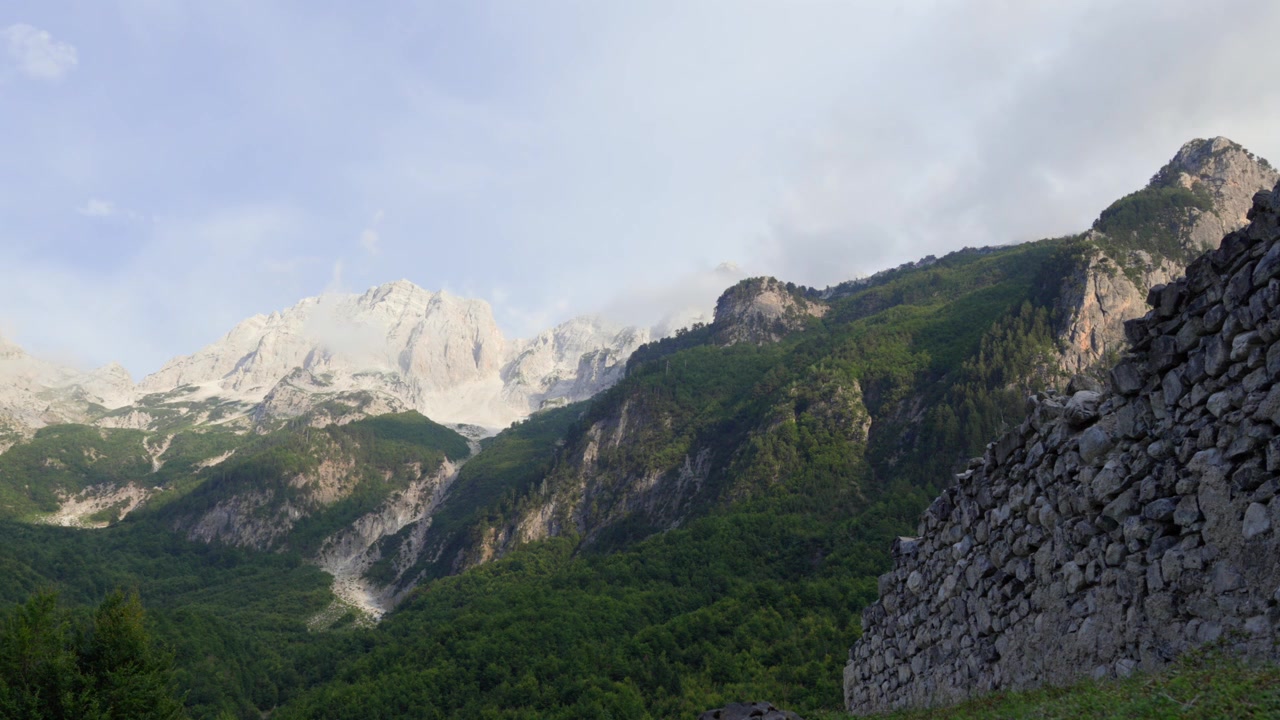 Valbona Vadisi Milli Parkı'nda, eski bir taş duvarın yanında, engebeli dağ zirveleri ve yemyeşil orman yamaçları üzerinde bulutlar nazikçe süzülüyor.